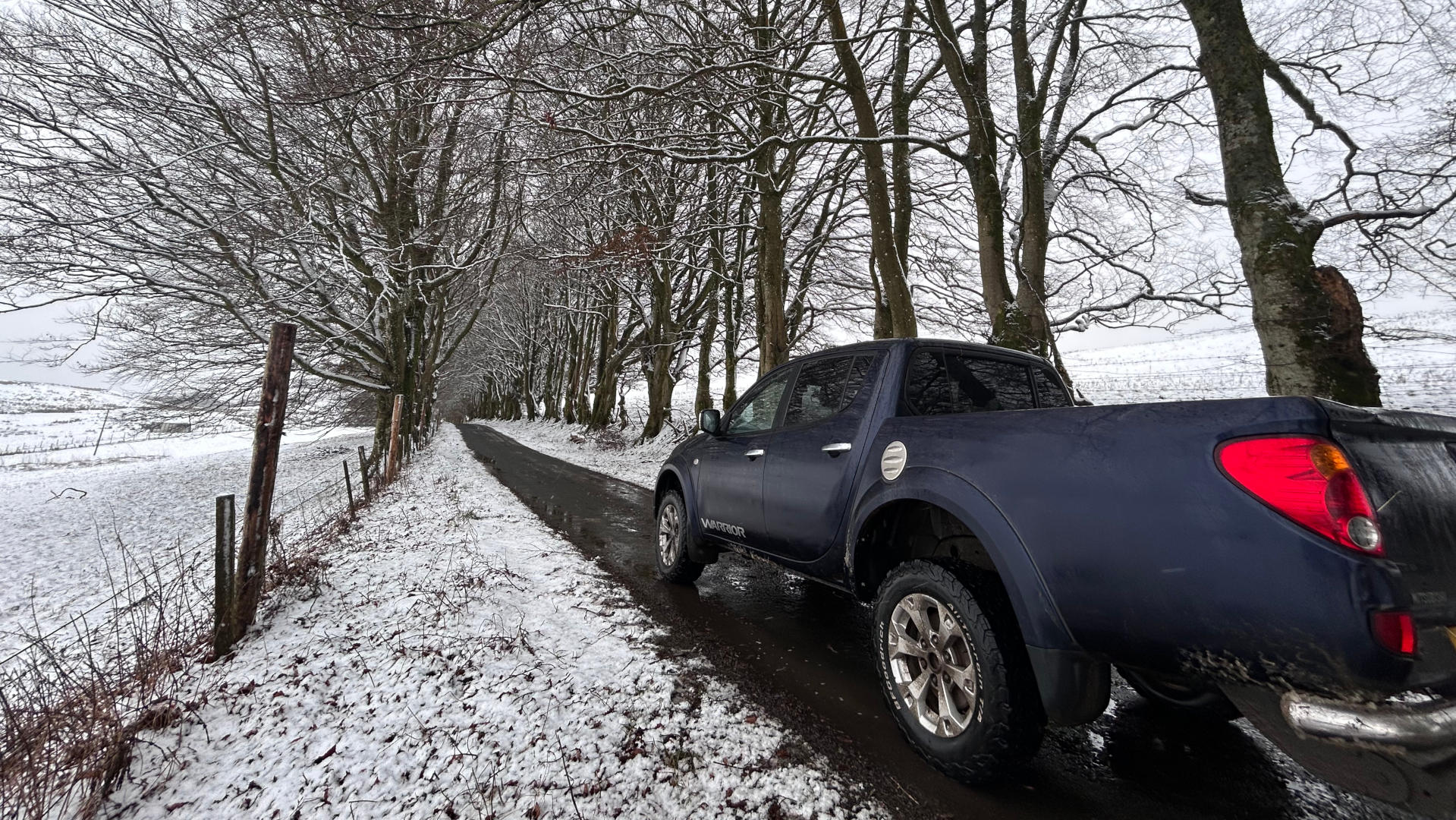 Dark-blue pickup truck parked on a snow-covered road that winds through a line of leafless, snow-dusted trees. The scene is quiet and peaceful, evoking a sense of winter solitude and the serenity of the countryside. The truck appears to be a Mitsubishi L200, based on the visible branding.