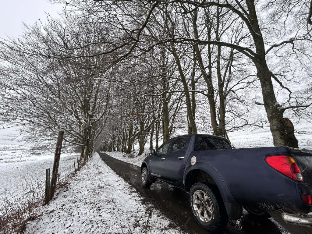 Dark-blue pickup truck parked on a snow-covered country road lined with bare, snow-dusted trees. The scene is peaceful and evokes a sense of quiet solitude in a wintry landscape. The road stretches into the distance, disappearing amidst the trees and the snow-covered fields.