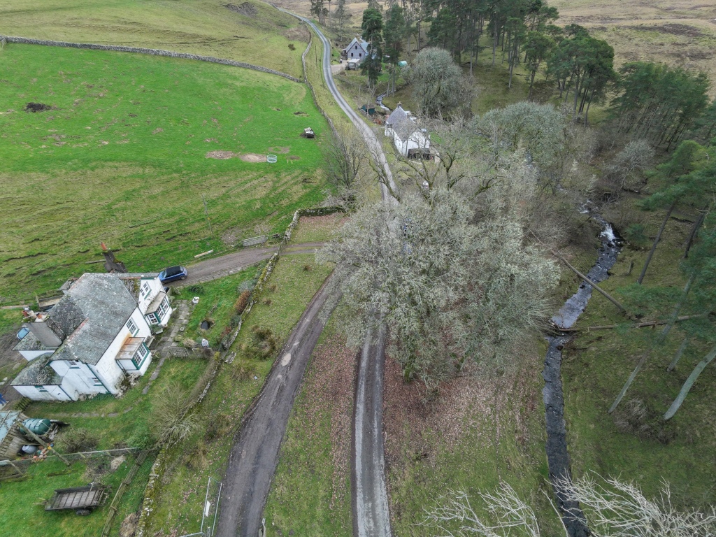 Aerial view of a rural landscape, featuring houses, a dirt road, fields, trees, and a stream. The green fields are bordered by stone walls. There is a house on the left side of the image with a blue car parked next to it. A dirt road runs through the center of the image, leading to other houses in the distance. A stream flows along the right side, bordered by trees.