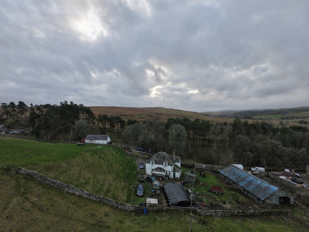 Rural landscape under a cloudy sky. In the foreground, there's a stone wall alongside a grassy hill. Beyond this, a small cluster of buildings, including a white house, a greenhouse, and a barn, are nestled amidst trees. The background features rolling hills extending into the distance under the overcast sky.