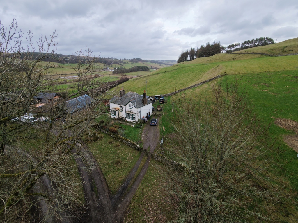 White farmhouse nestled amidst green, rolling hills under a cloudy sky. The house is a focal point, with a stone wall partially surrounding it and a dirt driveway leading up to it. Several vehicles are parked near the house. To the sides and in the foreground, there are bare trees without leaves, indicative of either late autumn or early spring. In the background, the landscape extends into the distance with a mix of fields, some wooded areas, and a ridge of trees lining the top of a hill.