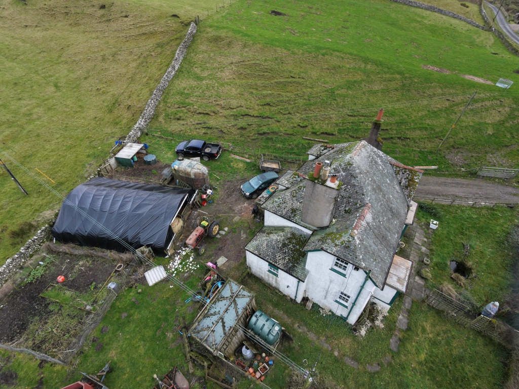 Aerial view of a rural farmhouse surrounded by patchwork green fields, stone walls, and muddy tracks. Weathered white buildings with mossy roofs sit beside sheds, tractors, and parked vehicles. Fences, water tanks, and footpaths reveal an active working farm within a quiet countryside landscape under overcast skies and rolling hills.