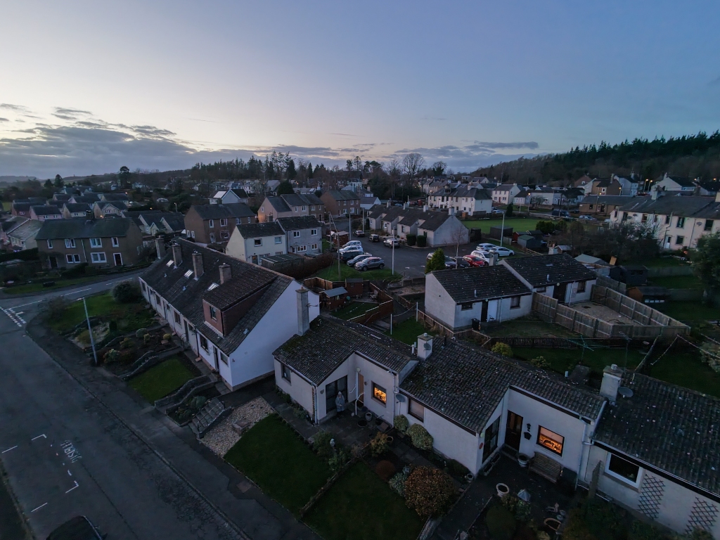 Aerial view of a quiet suburban neighborhood at dusk, rows of modest houses with tiled roofs lining curving streets. Warm lights glow through windows, gardens and fences define plots, parked cars fill a small lot, and wooded hills frame the settlement beneath a calm blue evening sky, overhead perspective tranquil.