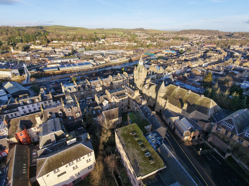 Aerial view of a historic town centre with tightly packed stone buildings, slate roofs, and narrow streets. A prominent clock tower rises above surrounding blocks, while a river and industrial area run behind. Sunlit hills, trees, and neighbourhood's spread outward, creating a layered urban landscape under clear skies today now.