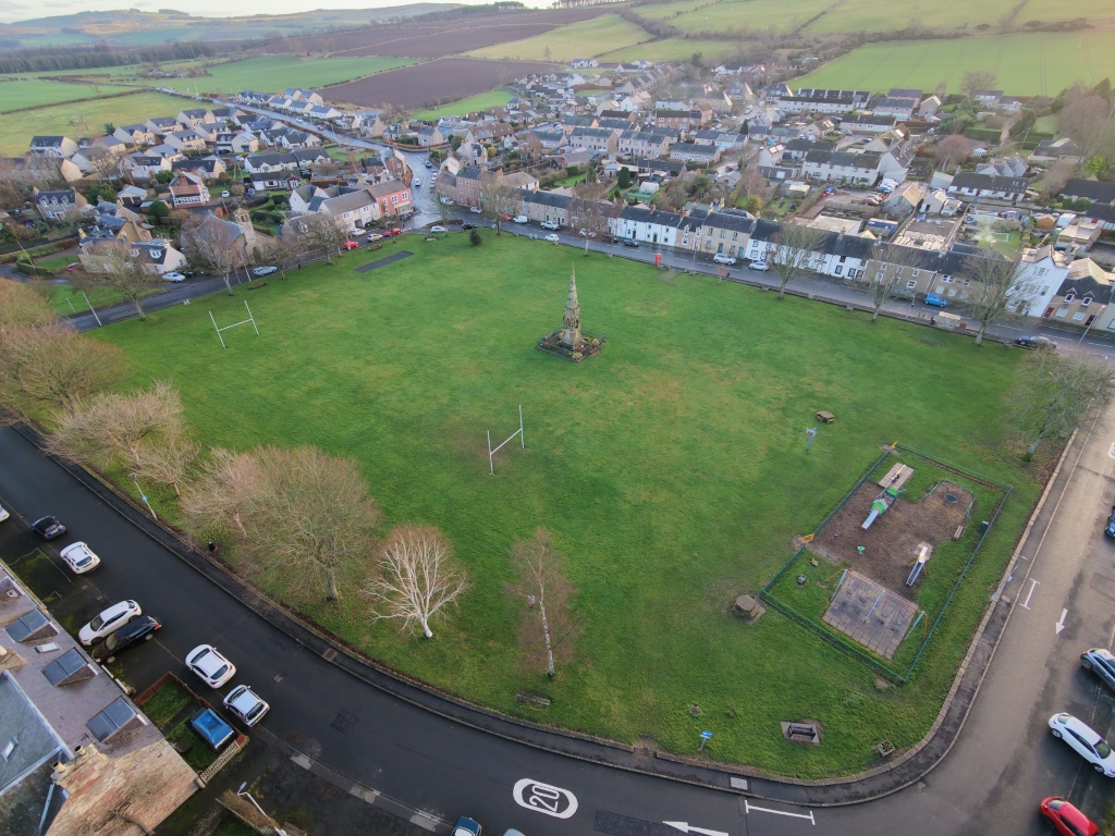 An aerial view shows a small town surrounding a large green common. A central stone monument stands amid open grass, with rugby goalposts and a fenced playground nearby. Rows of houses, parked cars, roads, and rolling farmland extend outward, under daylight. Trees line the edges, suggesting a quiet rural community.