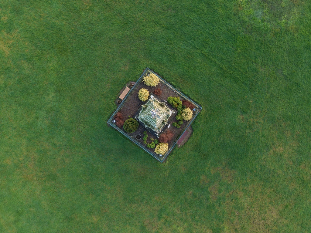 A top-down aerial view shows a small, square fenced monument set within a vast green lawn. The central stone structure is surrounded by shrubs and small trees arranged symmetrically. Benches line the enclosure, while uninterrupted grass stretches outward in all directions, emphasising isolation and geometric order.
