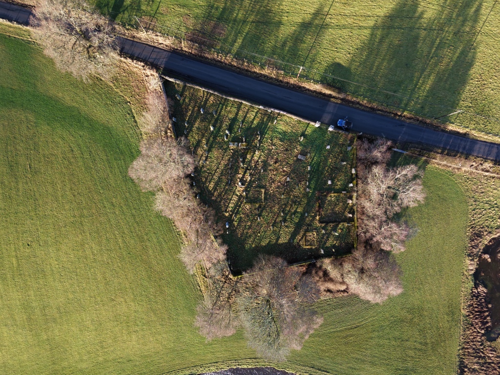 An aerial view shows a small, rectangular graveyard bordered by low stone walls and trees, set within open farmland. Gravestones cast long shadows across the grass, suggesting low sunlight. A narrow road runs along the top edge, with a parked car nearby, emphasising the rural, isolated setting.