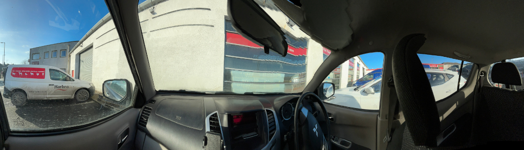 Panoramic view from inside a car, looking out at a building that appears to be a car repair shop or similar. A Harbro van is parked outside. Other cars are visible in the background, partially obscured by the vehicle the photo is taken from. The sky is clear and sunny.