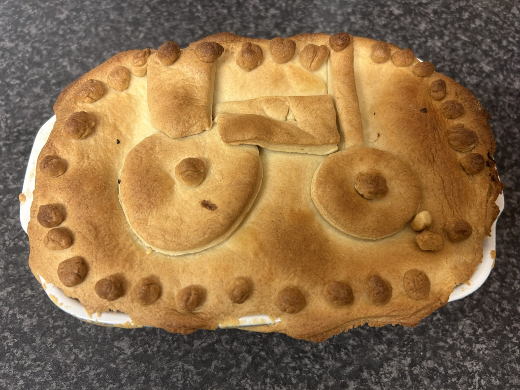 Baked pie with a pastry top designed to look like a tractor. The tractor design is formed using pieces of pastry to create the body, wheels, and even small details. The pie crust is golden brown and appears to be baked to a crisp.