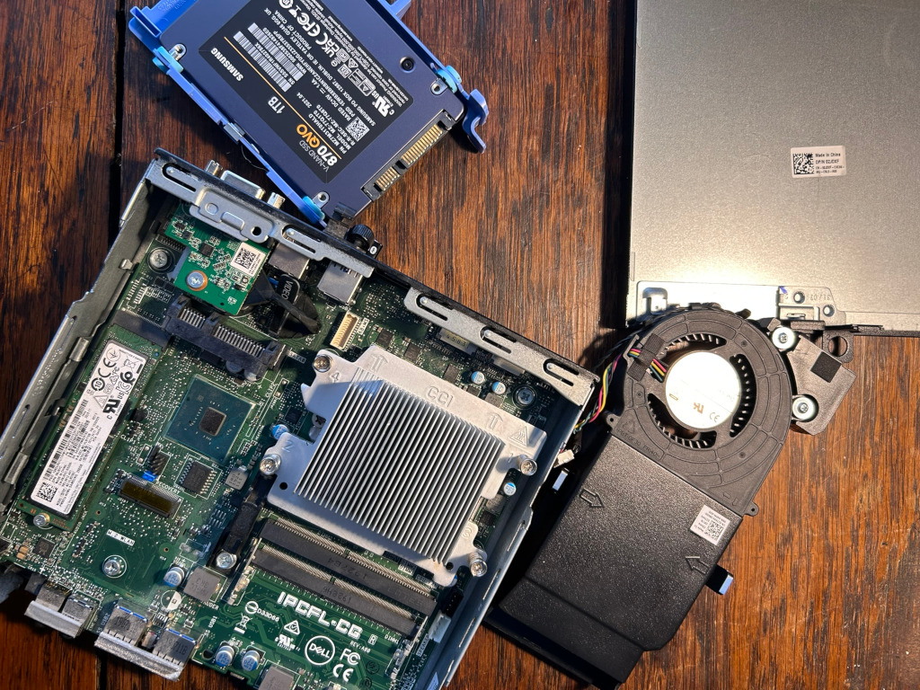 Disassembled components of a small form factor computer. The parts are spread out on a dark wooden surface. Visible components include: a motherboard, a solid-state drive (SSD) labelled Samsung 1TB 870 QVO a cooling fan assembly, and what appears to be a screen or display panel. The overall impression is one of repair or upgrade work in progress. There's no overt emotional expression or narrative, the image is purely descriptive of disassembled computer hardware. The moral is neutral; it simply documents a technical process.