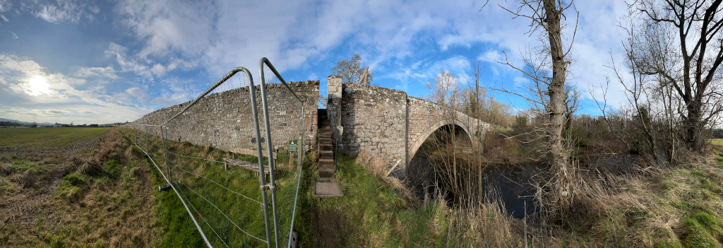 Panoramic view of an old stone bridge spanning a river. The bridge is partially enclosed by a low stone wall, and a temporary metal fence is partially visible, suggesting ongoing maintenance or repair work. The surrounding landscape is largely flat farmland, with bare winter trees lining the riverbank. The sky is partly cloudy with bright sunshine.