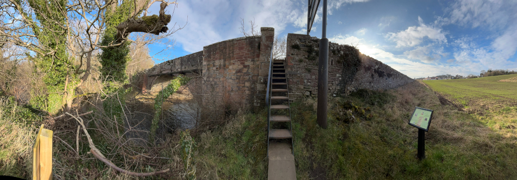 Panoramic view of an old stone bridge over a waterway. A metal staircase leads up to the bridge from the right. On the right of the stairs, a small informational sign is visible, and to the left, the bridge spans over what appears to be a dry or low water level. The area is overgrown with vegetation, and a field stretches in the background. The sky is partly cloudy.