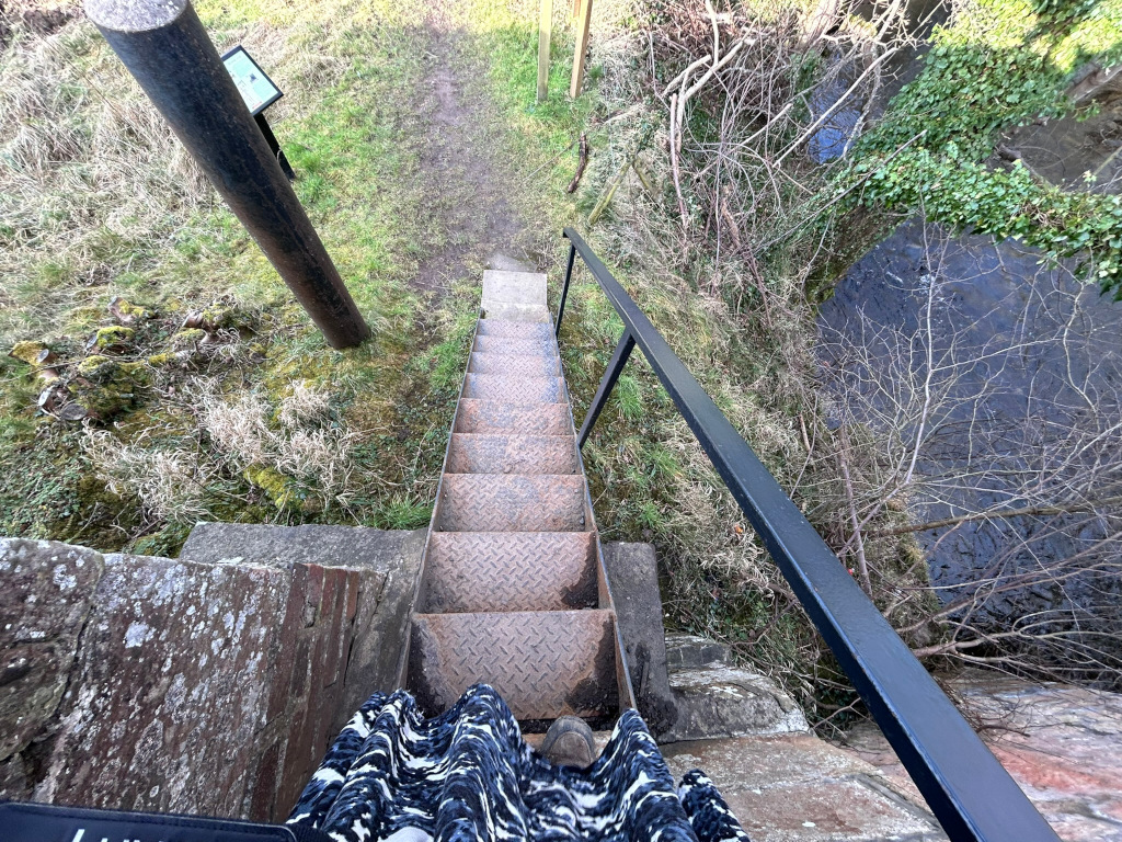 Leonie standing at the top of a metal staircase that descends towards a small stream. The stairs are rusty and appear somewhat old and worn, and are bordered by a black handrail. The surrounding area is overgrown with grass and vegetation. A portion of Leonie's patterned skirt is visible at the bottom of the frame. To the left, there's a weathered stone wall and a wooden post with a small sign. The overall impression is one of a rustic, perhaps somewhat secluded, outdoor location.