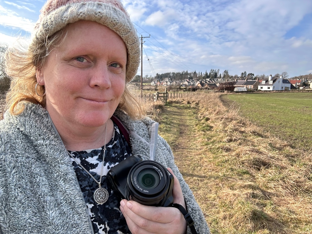 Middle-aged woman (Leonie) with blonde hair wearing a knit hat and a grey fleece jacket. She is holding a compact digital camera and is standing on a path next to a field.  In the background, there's a small village or collection of houses under a bright, sunny sky. The overall impression is one of a leisurely outdoor excursion, perhaps a casual photography outing in a rural setting.