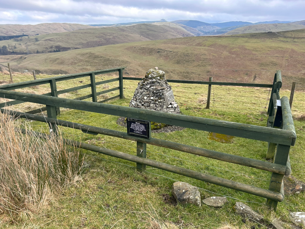 Cairn, a man-made pile of stones, enclosed by a dark green wooden fence. A small plaque is affixed to the cairn. The background is a sweeping vista of rolling green hills under a cloudy sky. The scene appears to be rural and possibly in a mountainous region.