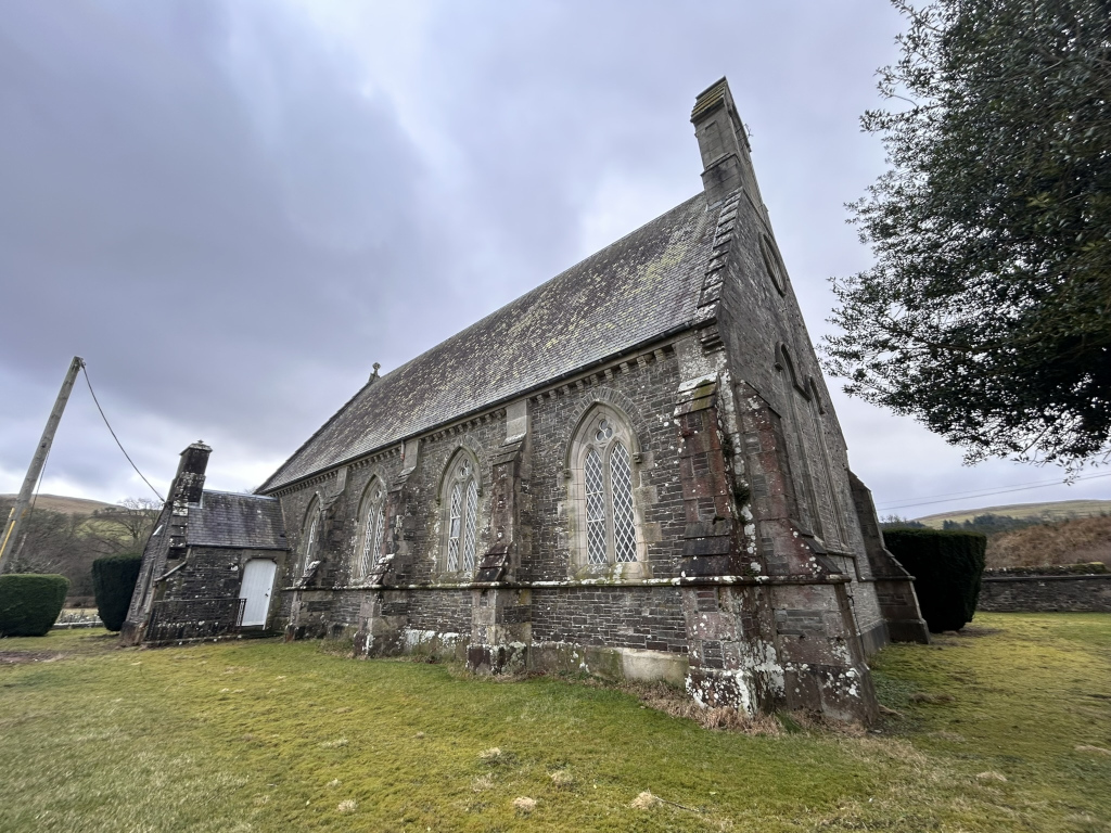 Side profile of a stone church building under a cloudy sky. The church is made of dark grey stone, with a steeply pitched slate roof and several tall, narrow, arched windows. The structure is somewhat aged and shows signs of weathering. A small, attached structure is visible at the rear. The foreground consists of short, grassy land, and part of a tree is visible in the upper right corner, framing the church. The overall impression is one of quiet solitude and aged architecture in a rural setting.