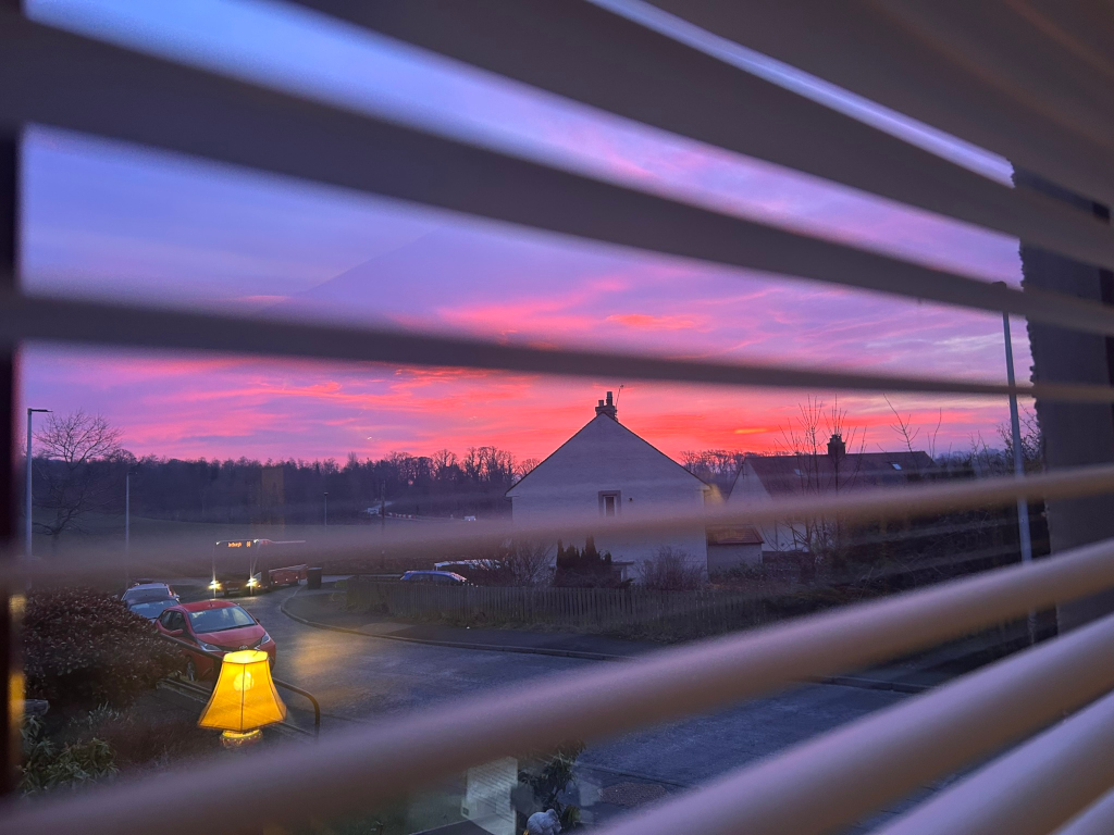 A vibrant sunset viewed through the slightly ajar slats of a window blind. The setting sun paints the sky in shades of pink, orange, and purple. In the foreground, a street scene is visible, featuring houses, cars, and a lamp. The overall mood is peaceful and serene.