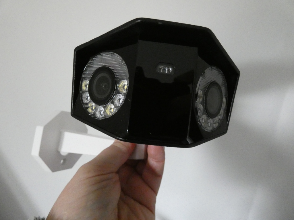 Black security camera with multiple infrared LEDs surrounding the lenses. It's mounted on a white bracket, Leonie's hand is holding the camera and bracket. The background is a plain light-grey wall.