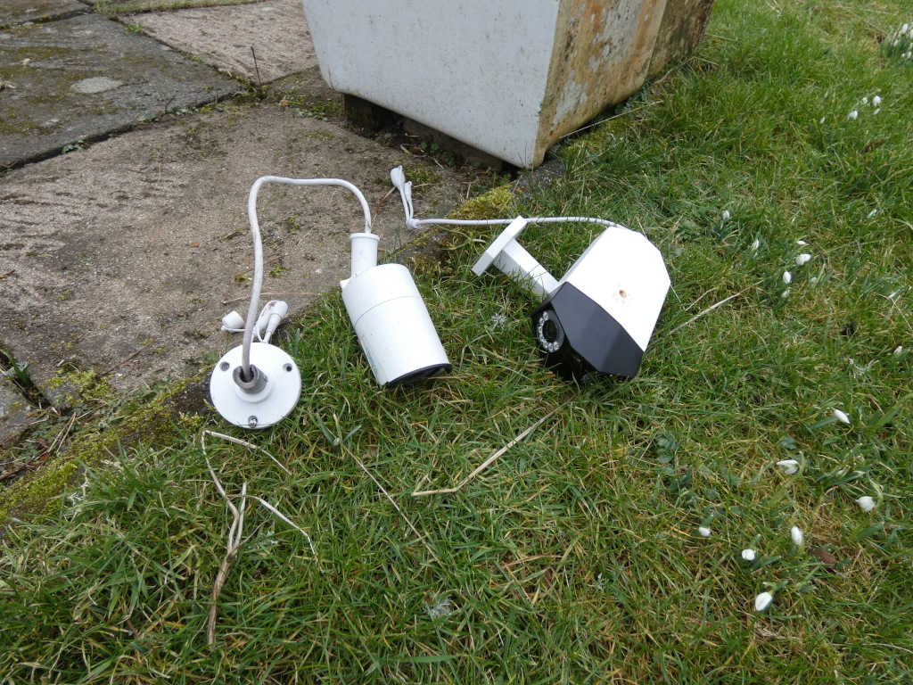 Two discarded or abandoned white and black security cameras lying in the grass next to a paved area. One camera is cylindrical and the other is angular. Both appear to be detached from their mounts and have wires trailing from them. The scene suggests removal of a security system.