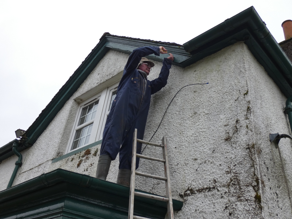 Charlie standing on a ladder against the corner of a building. He is wearing a dark blue jumpsuit and appears to be working on the building's exterior, possibly performing maintenance or repair work on the gutter or wiring. The building is an older structure with textured white walls and dark green trim. The overall scene is one of everyday maintenance or repair work.