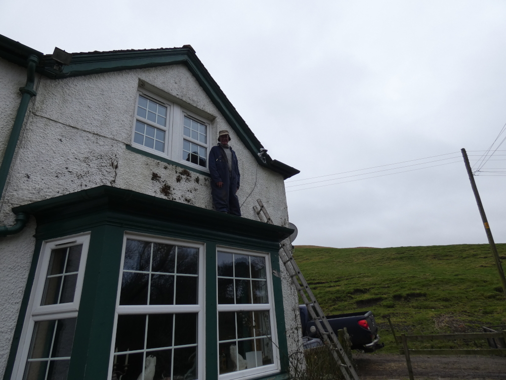 Charlie in overalls standing on the roof-line of a house, just above a bay window. He appears to be a worker, possibly on a repair or maintenance job. A ladder leans against the house, suggesting his work. The house is white with dark green window frames and trim and is situated in a rural setting with a grassy hill visible in the background. The sky is overcast.