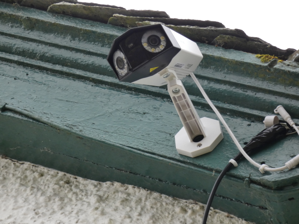 Security camera mounted on the exterior of a building. The camera is black and white, with a rectangular shape. It is mounted on a white bracket, which is attached to the building's green painted eaves. The wires connecting the camera are visible. The building's exterior is a combination of painted wood and stucco. The overall impression is one of security and surveillance.