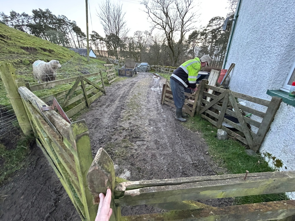 A muddy driveway leading to a farm house. Charlie in a high-visibility jacket is working on a section of wooden fencing near the house. A sheep is visible in a fenced area on the left side of the driveway. The overall setting appears rural and somewhat rustic. The focus of the image is from a low angle, giving the viewer a perspective of looking up along the driveway and towards the house.