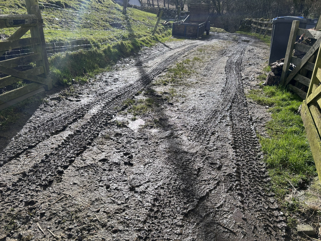 Muddy track or lane, deeply rutted by vehicle tires. The track leads from a wooden gate on the left to a small cart or trailer parked further up. The surrounding area is rural, with green grass and a stone wall visible in the background. The overall impression is of a farm track after recent rain.