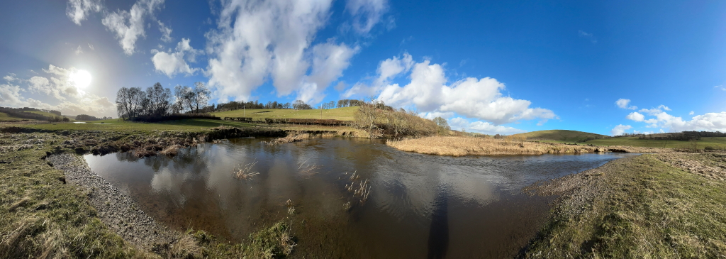 Panoramic view of a tranquil river scene on a sunny day. The river meanders through a gently rolling landscape of grassy fields and sparse woodland under a bright blue sky dotted with fluffy white clouds. The sunlight is bright, casting reflections on the calm water.