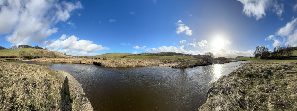Panoramic view of a serene river scene on a bright, sunny day. The river meanders through a gently rolling landscape of grassy fields and sparsely wooded hills under a mostly clear blue sky with scattered fluffy white clouds. The sun is visible in the sky. The overall impression is one of peace and tranquillity.