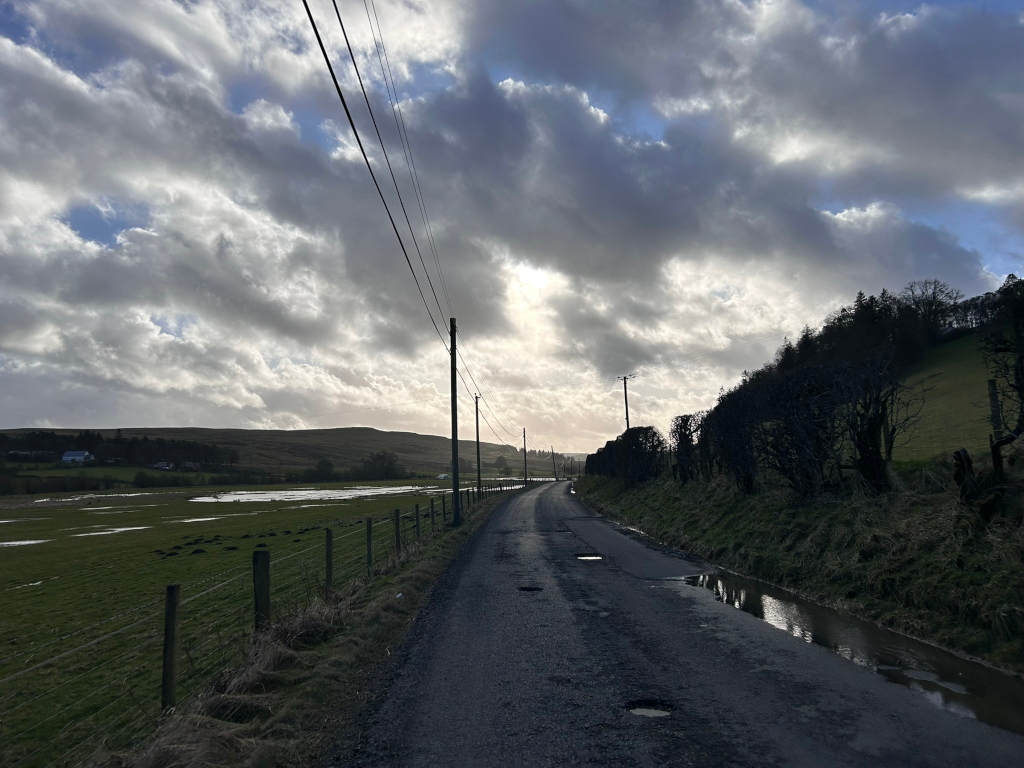 Rural road stretching towards a distant hilly landscape under a dramatic, partly cloudy sky. The road is relatively narrow and shows signs of wear, with some potholes visible. The roadside features a fence separating the road from a grassy field with some puddles, suggesting recent rain. In the distance, low hills and a line of trees are visible. The overall mood is somewhat bleak yet peaceful, with a contrast between the dark road and the lighter sky.