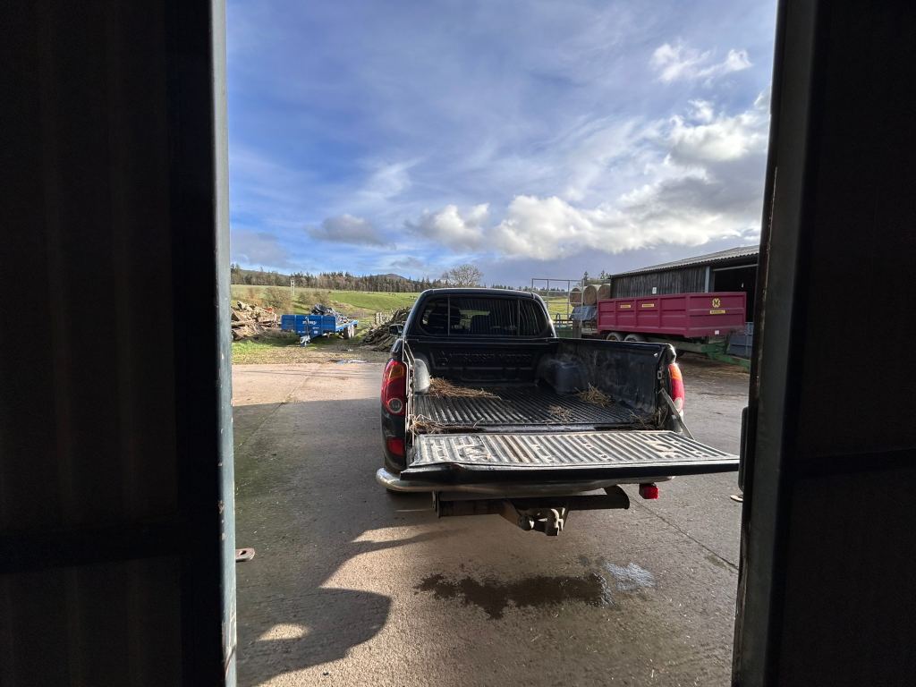 Dark-coloured pickup truck with its tailgate down, parked in a farmyard. The truck bed contains some hay or straw. The background includes farm buildings, agricultural machinery, and a partly cloudy sky. The view is framed by a partially open doorway or gate.
