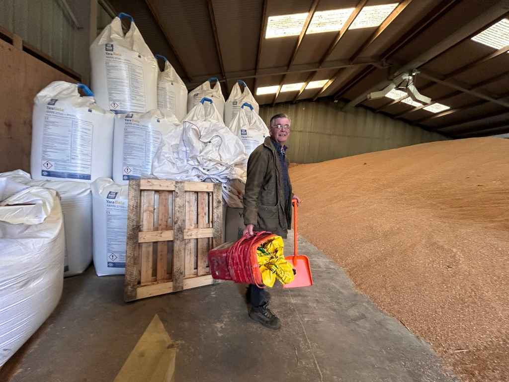 Charlie standing in a large agricultural building, likely a grain store. He's holding a red bucket and an orange shovel. Behind him is a large pile of grain or similar agricultural product, and several large sacks of fertiliser are stacked near him. The overall impression is one of agricultural work or storage.