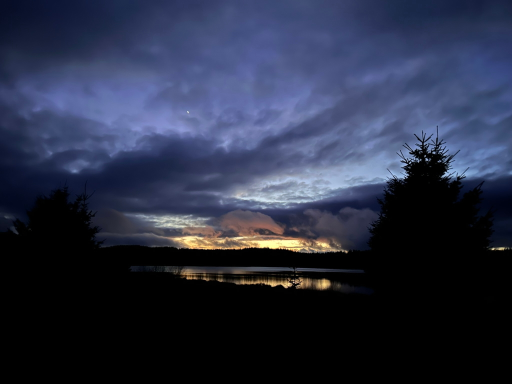 Serene twilight scene over a body of water. The sky is dominated by a dramatic display of dark, brooding clouds, punctuated by a sliver of warm, colourful light near the horizon suggesting either sunrise or sunset. Silhouetted evergreen trees flank the scene, emphasising the darkness and stillness of the landscape. A single, small light, possibly a star or planet, is visible in the upper portion of the frame, adding to the feeling of vastness and tranquillity. The overall mood is peaceful but slightly melancholic, with a contrast between the darkness of the land and the vibrant yet subdued colours of the sky.