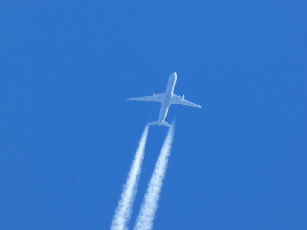 White passenger jet aeroplane flying high in a clear, bright blue sky. A contrail, the vapour trail left by the plane's engines, is clearly visible extending down from the aircraft. The overall impression is one of vastness and travel.