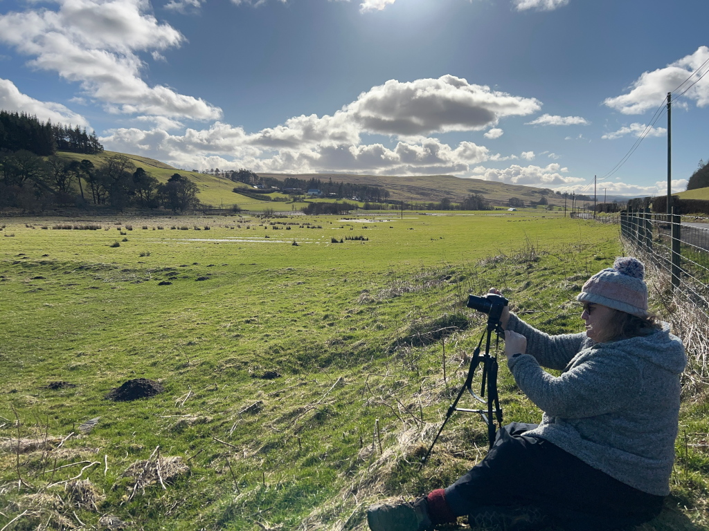 Leonie wearing a grey sweater and a pink beanie, sitting in a grassy field and taking a photograph with a camera mounted on a tripod. The background features a pastoral landscape with rolling green hills, a partly cloudy sky, and a road. The overall impression is one of peaceful outdoor photography in a serene rural setting.