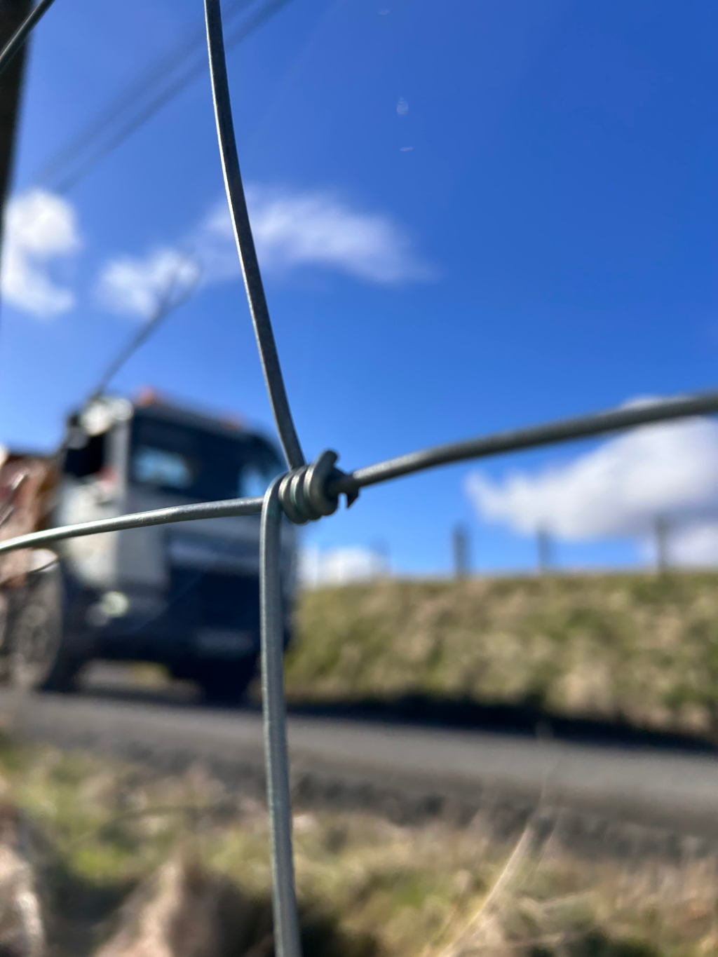 Close-up view of a wire fence knot, with a blurred background of a truck driving on a road in a rural setting under a bright blue sky. The focus is sharply on the wire knot and the background is out of focus, suggesting a deliberate artistic choice to highlight the texture and detail of the fence. The overall impression is one of a simple, everyday scene observed through a specific, limited perspective.
