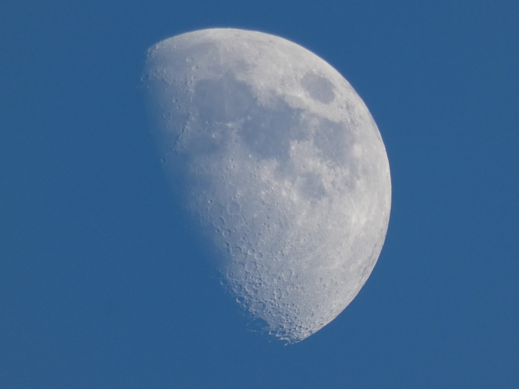 Gibbous moon against a clear blue sky. The moon is approximately three-quarters full, displaying significant surface detail including craters and maria (darker, smoother areas). The overall impression is one of serenity and vastness.