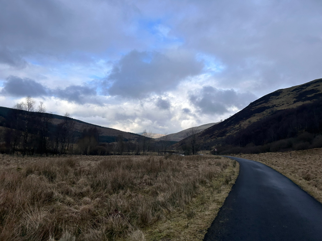 Paved road curving gently through a valley landscape. The road is flanked by dry, brown grass and leads towards distant, low hills under a cloudy sky. The hills are sparsely covered with dark-coloured vegetation. The overall mood is serene and somewhat desolate.  There's a pale patch of blue sky visible between the clouds, hinting at a break in the weather.