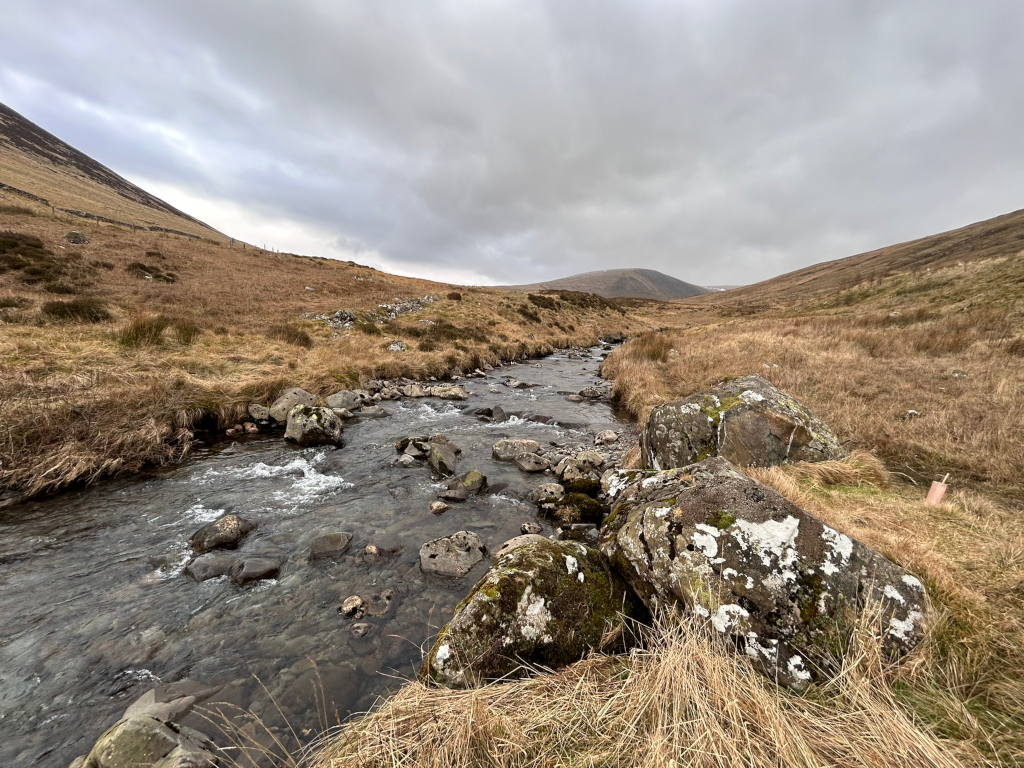 Serene landscape featuring a shallow stream meandering through a valley. The stream is filled with smooth, dark rocks, and its water appears relatively clear. The valley is characterised by dry, light brown grasslands, that slope gently upwards to meet the hills. The sky is overcast, with a muted, grey tone dominating the scene.