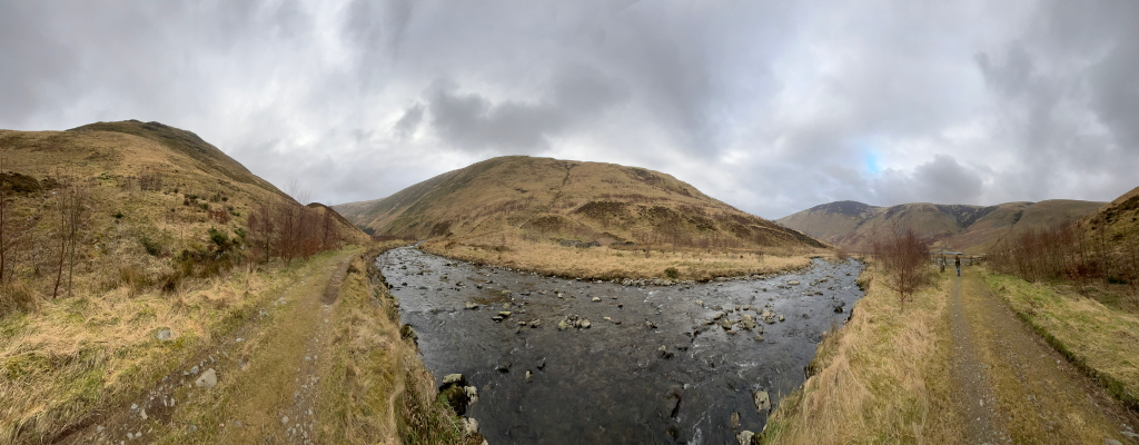 Panoramic view of a valley scene. A river meanders through the valley floor, flanked by grassy, brown hillsides under a cloudy sky. Two dirt paths/roads are visible, one on each side of the river, Charlie is walking on one of the paths in the distance. The overall mood is serene and somewhat desolate.