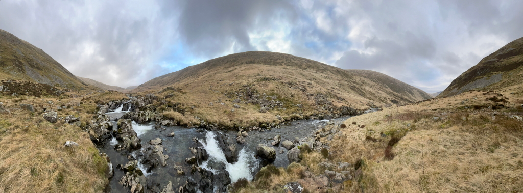 Panoramic view of a mountainous landscape. A river flows through the valley, cascading over rocks in a small waterfall. The hills are covered in dry, light brown grass, and the sky is mostly cloudy, with patches of blue visible. The overall impression is one of wild, remote beauty, possibly in a highland or upland area.