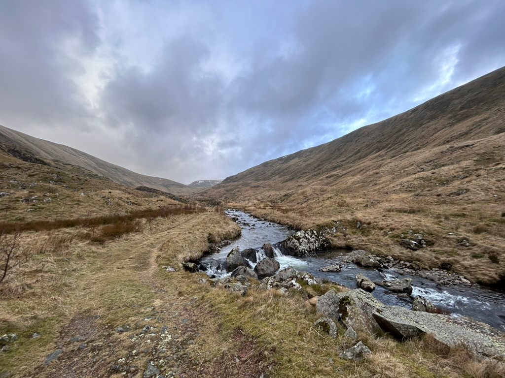 Tranquil mountain stream flowing through a narrow valley.  The valley slopes are covered in dry, light brown grass, typical of a temperate upland or highland region. A faint trail or path is visible along the valley floor near the stream. The sky is heavily clouded, giving the scene a somewhat sombre or muted tone. The overall impression is one of serene, yet somewhat harsh, natural beauty.