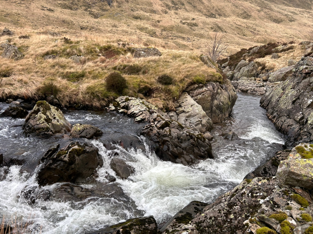 Mountain stream cascading over dark rocks. The water is white and frothy, indicating a relatively fast current. The stream flows through a narrow channel carved into the landscape. The surrounding land is sparsely vegetated with dry, brownish grass and some patches of darker green