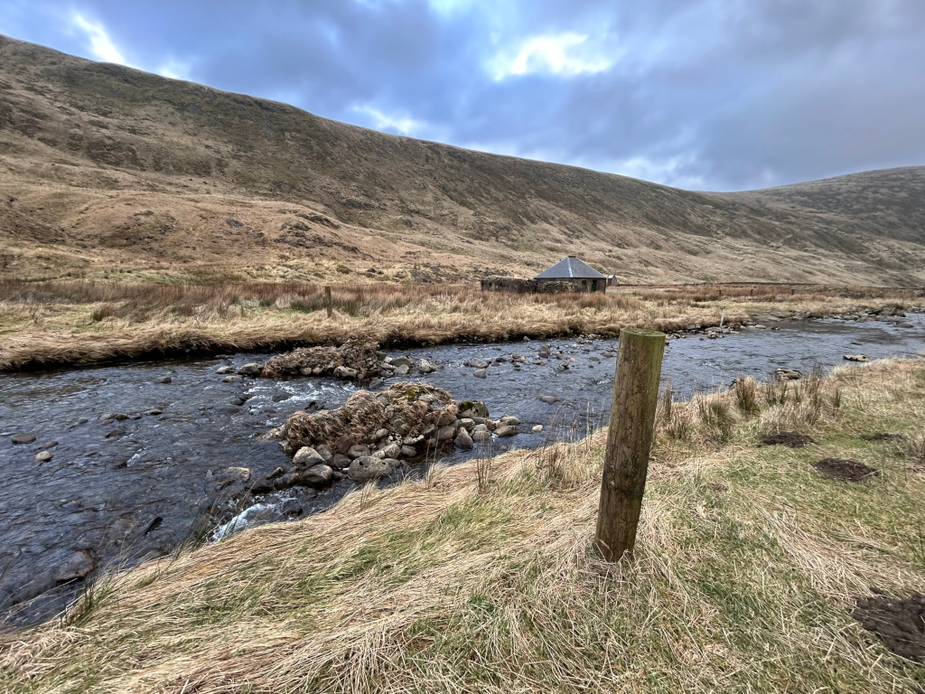 Calm river flowing through a sparsely vegetated valley. A small, round stone bothy is visible in the middle ground against the backdrop of rolling, brown hills under a cloudy sky. A wooden post stands near the riverbank in the foreground.
