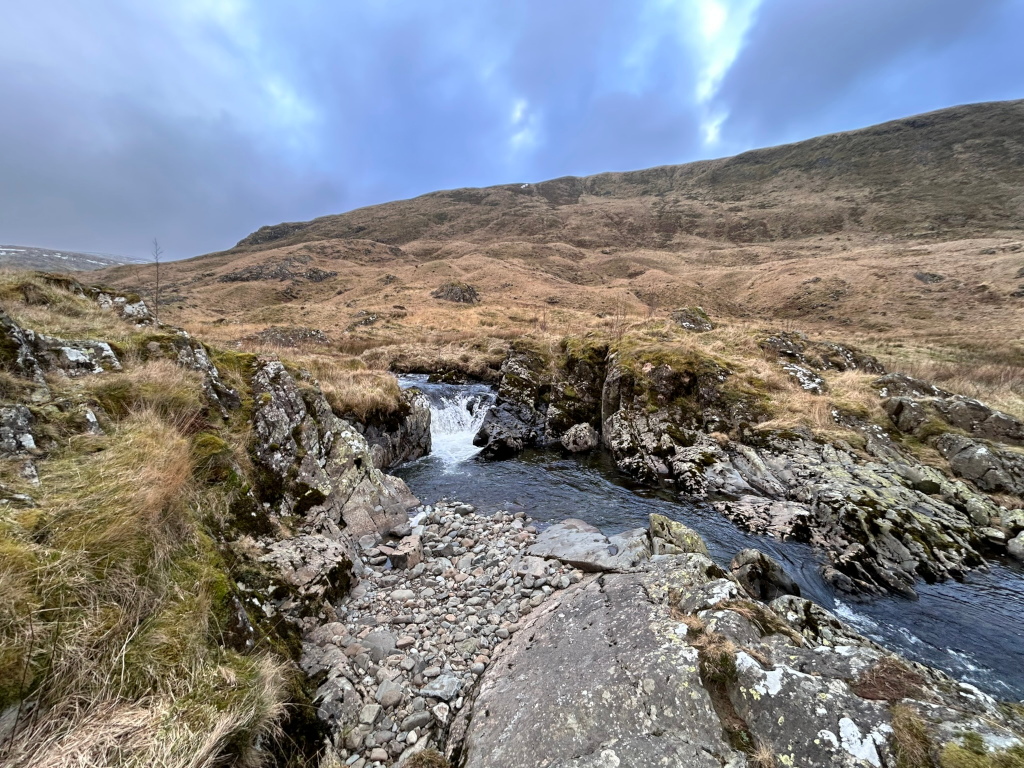 Small waterfall cascading down rocky terrain into a stream. The stream flows through a rocky, sparsely vegetated landscape, with a hillside rising in the background under a somewhat cloudy sky. The overall scene is one of a wild, natural, and somewhat desolate beauty.