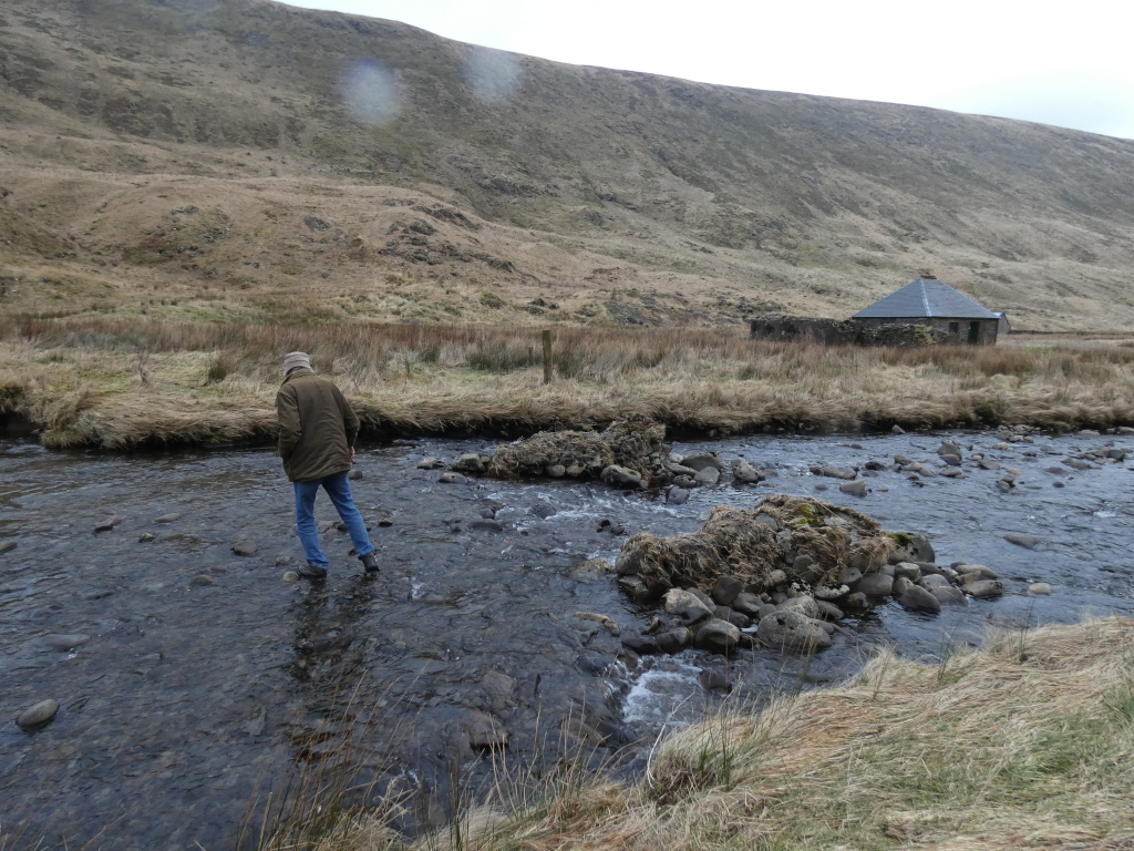 Charlie walking across a shallow river. The river bed is rocky and the water is relatively clear. In the background, there is a small stone bothy and a large hill. The overall scene is rural and somewhat desolate. The man is walking across the river on stepping stones, which are rocks sticking out of the shallow water. The sky is overcast, and the overall colour palette is muted.