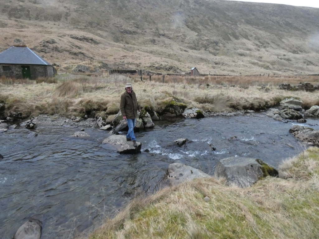 Charlie in a brown jacket and blue jeans crossing a shallow stream. He is stepping on rocks in the middle of the stream. The stream is situated in a rural, mountainous area, with dry, brown grass and low stone bothy is visible in the background. The overall atmosphere is serene and somewhat solitary.