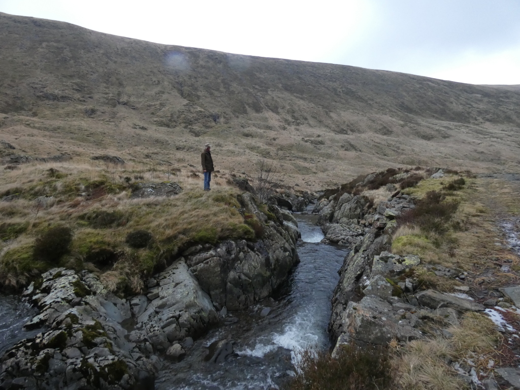 Charlie standing on a rocky outcrop overlooking a small stream flowing through a valley. The valley has a barren, mountainous landscape with brown and grey hues. The overall mood is calm and somewhat isolated. Charlie appears small in comparison to the vastness of the landscape.