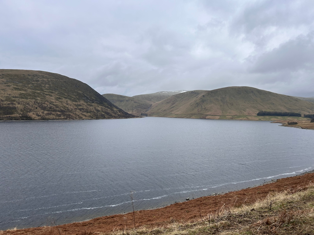 Serene landscape featuring a calm body of water, possibly a lake or reservoir, nestled between gently rolling hills. The hills are sparsely vegetated, appearing somewhat barren, with patches of brown and green. The sky is overcast, a muted grey, adding to the overall tranquil, yet somewhat melancholic atmosphere. The water shows light ripples, suggesting a subtle breeze. The overall impression is one of quiet solitude and natural beauty in a somewhat remote location.
